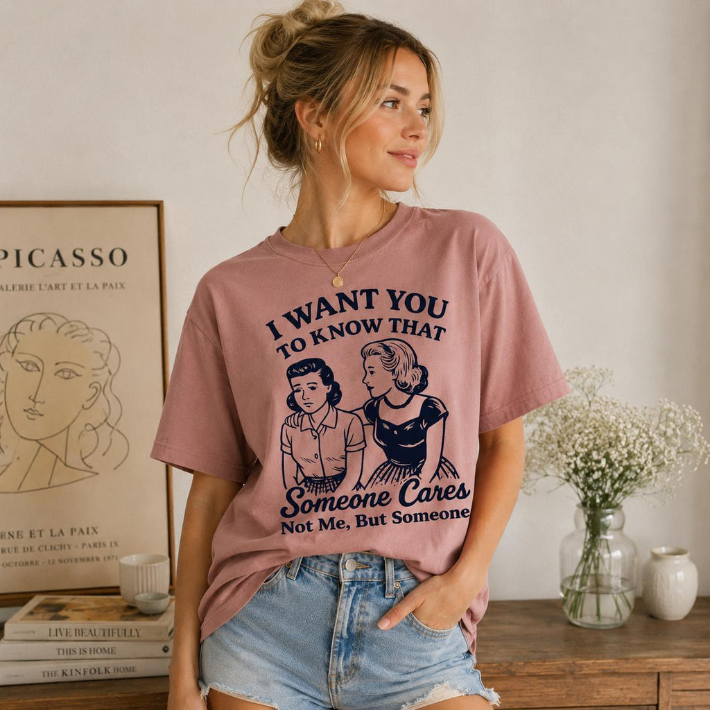 Woman wearing a mauve graphic tee with a retro illustration and funny text, near flowers and art.