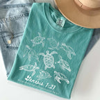A teal garment-dyed t-shirt featuring white sea turtle illustrations and Genesis 1:21 scripture on jeans with a hat.