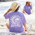 Violet graphic t-shirt with white sun and palm trees on the back, worn by a woman on a sandy beach with the ocean in the