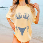 Woman wearing a pale yellow oversized cotton graphic t-shirt with a seashell bikini body illusion print while posing on a