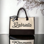 Custom name tote bag featuring a two-tone canvas body, black script name Gabriella, and a striped ribbon tie on handle.