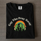 Black graphic t-shirt folded on wood surface featuring arched Gay The Pray Away text over three dancing frogs, rainbow, and