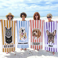 Four women holding personalized pet portrait beach towels with yellow, blue, red, and purple stripes on a sandy beach.
