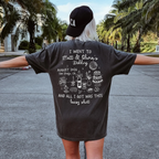 A woman wearing a charcoal gray funny wedding t shirt stands outside with arms outstretched near palm trees.