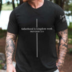 A tattooed man outdoors wearing a black t-shirt printed with a white cross and the phrase fatherhood is kingdom work proverbs