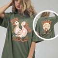 A woman wearing an olive green oversized t-shirt with a duck duck print on front and goose print on back.
