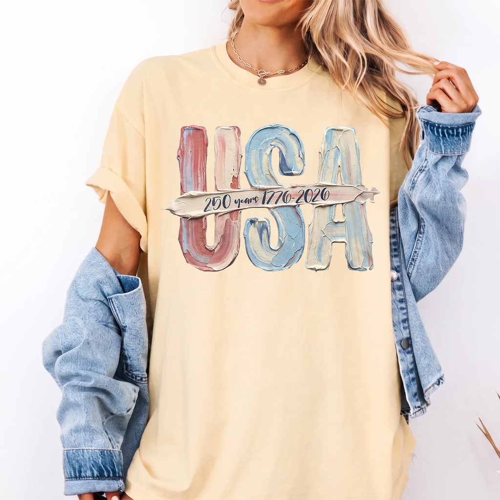 A woman wearing a yellow graphic tee with muted red, cream, and blue USA lettering and a denim jacket.