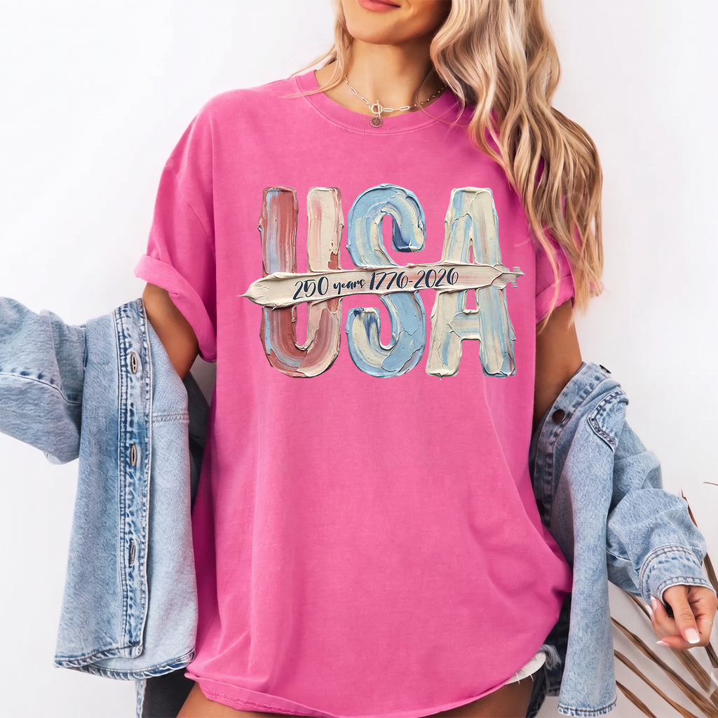 A woman in a pink garment-dyed graphic t-shirt with textured USA script and a denim jacket draped over her.