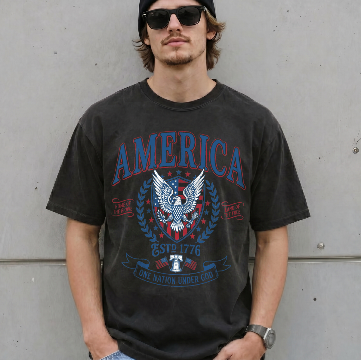 Man wearing a dark grey vintage America patriotic eagle graphic tee with sunglasses in front of a concrete wall