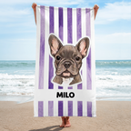 Purple striped personalized French Bulldog beach towel held up on a sunny beach against the ocean and sky.