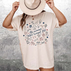 Woman wearing cream cotton graphic t-shirt featuring 4th of July doodle design and cowgirl hat against rustic metal