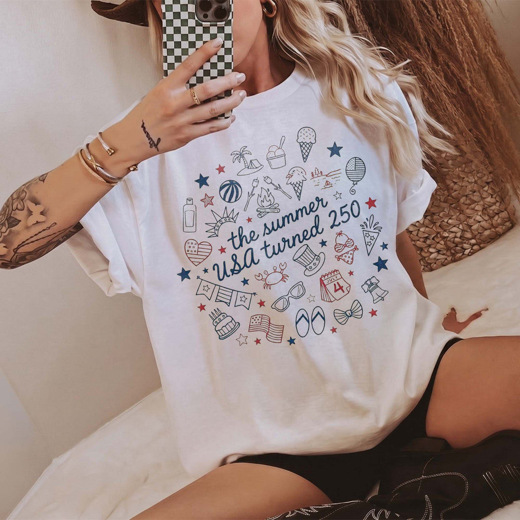Seated woman wearing white cotton graphic t-shirt with patriotic summer doodle print and black shorts.