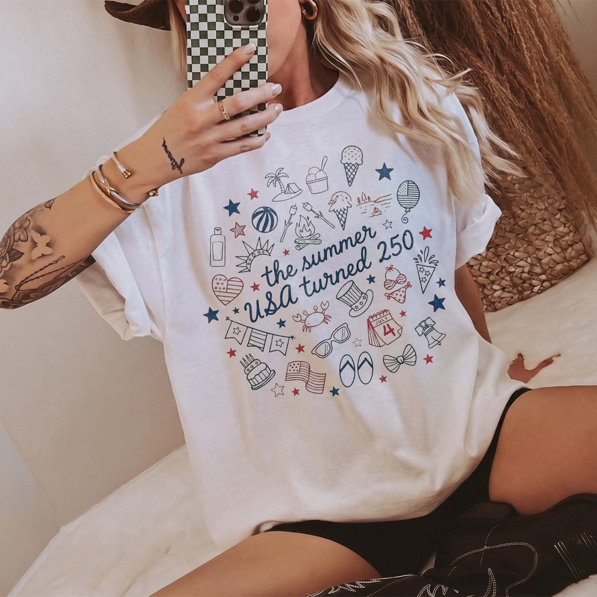 Seated woman wearing white cotton graphic t-shirt with patriotic summer doodle print and black shorts.