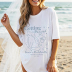 A woman wearing a white graphic t-shirt with a cat bride design and text near a beach holding a bridal veil.