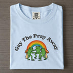Light blue graphic t-shirt folded on wood surface with arched Gay The Pray Away text, three dancing frogs, rainbow, and