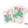 Ivory t-shirt featuring green alligator graphic with star sunglasses, Later and After While text, and stars on a white