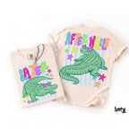 Ivory t-shirt featuring green alligator graphic with star sunglasses, Later and After While text, and stars on a white