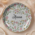 Round floral ceramic trinket dish on textured background featuring pink tulips, green leaves, gold rim, and the name Anna.