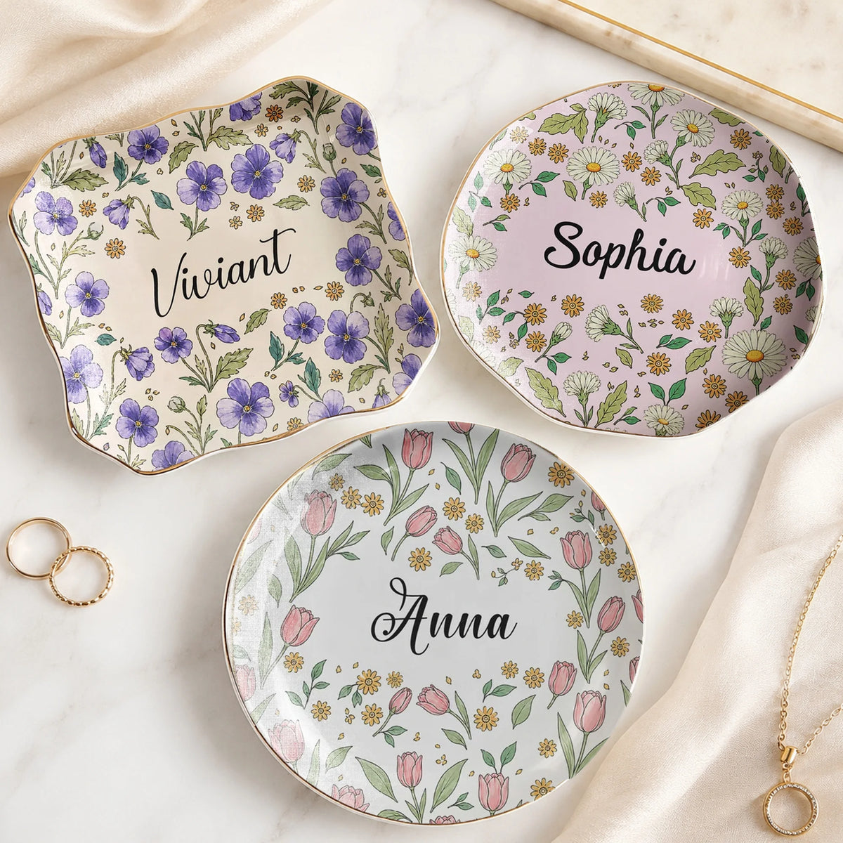 Three custom name jewelry trays in round, square, and wavy shapes featuring botanical floral designs and gold rims.