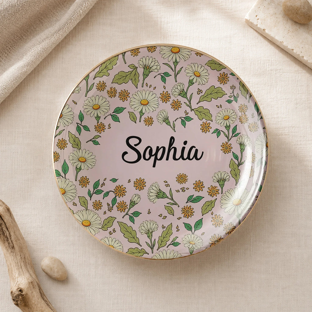 Round floral ceramic trinket dish on neutral fabric with pink background, white daisies, gold rim, and the name Sophia.