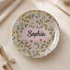 Round floral ceramic trinket dish on neutral fabric with pink background, white daisies, gold rim, and the name Sophia.