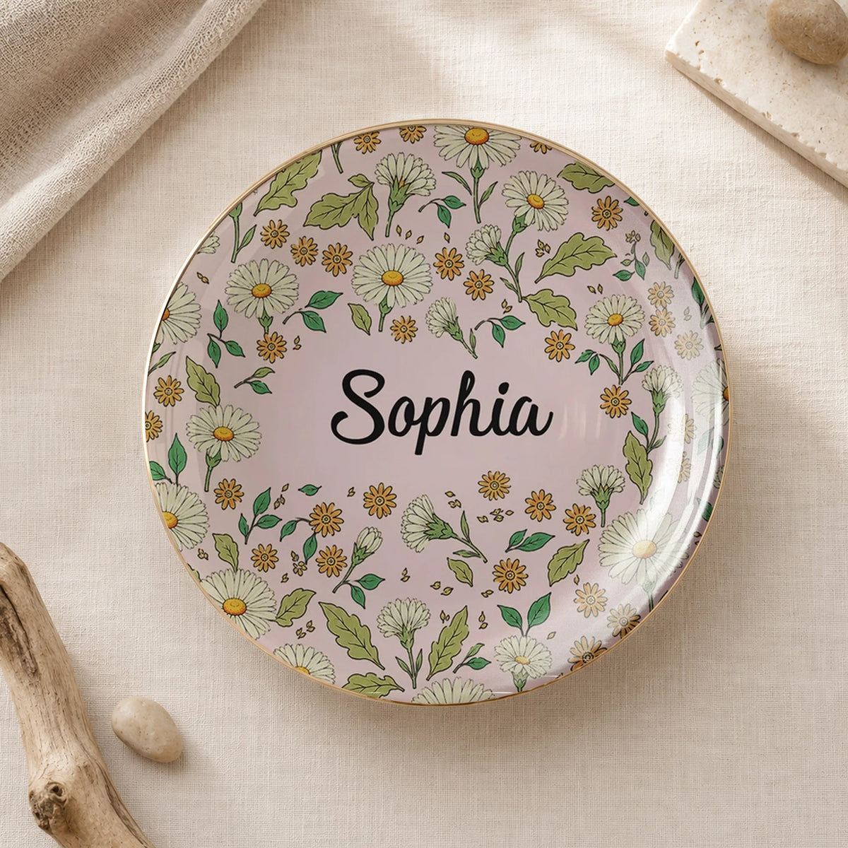 Round floral ceramic trinket dish on neutral fabric with pink background, white daisies, gold rim, and the name Sophia.