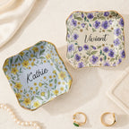 Two wavy square ceramic ring dishes with purple and yellow botanical birth flower prints and custom names.