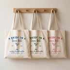 Three natural canvas tote bags hanging on wooden hooks, featuring blue, green, and pink martini graphics for bachelorette