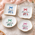 Four personalized white porcelain cheerleading trinket trays with gold rims and watercolor megaphone and bow designs.