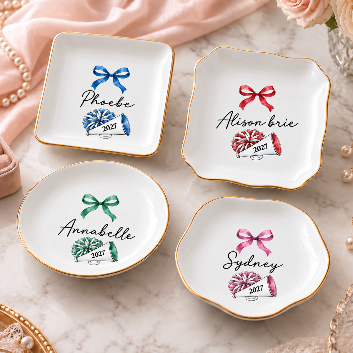 Four personalized white porcelain cheerleading trinket trays with gold rims and watercolor megaphone and bow designs.