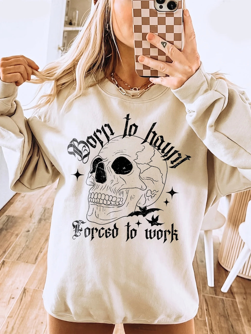 A blonde woman wearing a light beige graphic sweatshirt featuring a distressed skull, bats, and Born to haunt Forced to work
