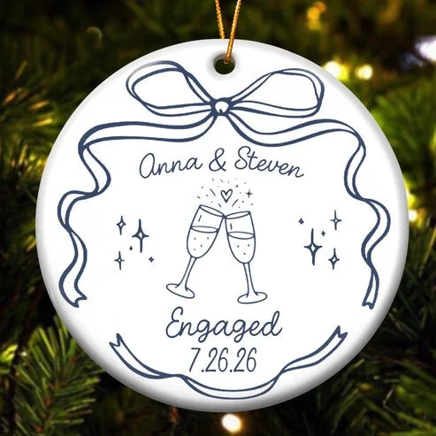 A round white ceramic ornament featuring clinking champagne glasses graphic, names, and date hanging against pine tree