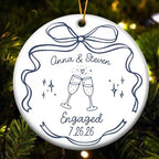 A round white ceramic ornament featuring clinking champagne glasses graphic, names, and date hanging against pine tree