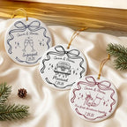 Three round ceramic holiday ornaments displaying car, glasses, and pinky promise designs resting on a silky beige fabric