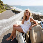 Bride wearing an off-white Bride and Bridesmaid Ruffle Pajama Set and veil sitting in a convertible by the sea.