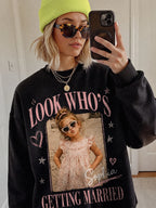 A person in a black custom photo t-shirt wearing a yellow beanie, tortoiseshell sunglasses, and taking a mirror selfie.